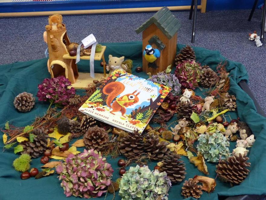 A vibrant autumn-themed display featuring the children's book 'Leaf Thief' surrounded by pinecones, dried flowers, chestnuts, and small woodland animal figurines on a green cloth.