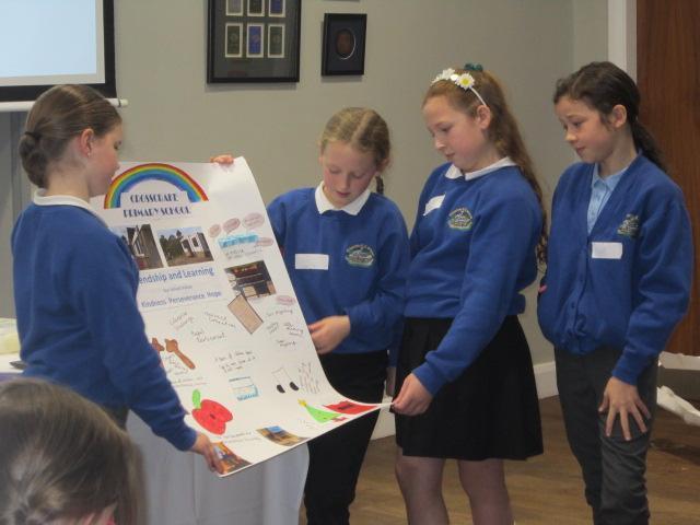 Four primary schoolgirls in blue uniforms present a large, colourful project board titled 