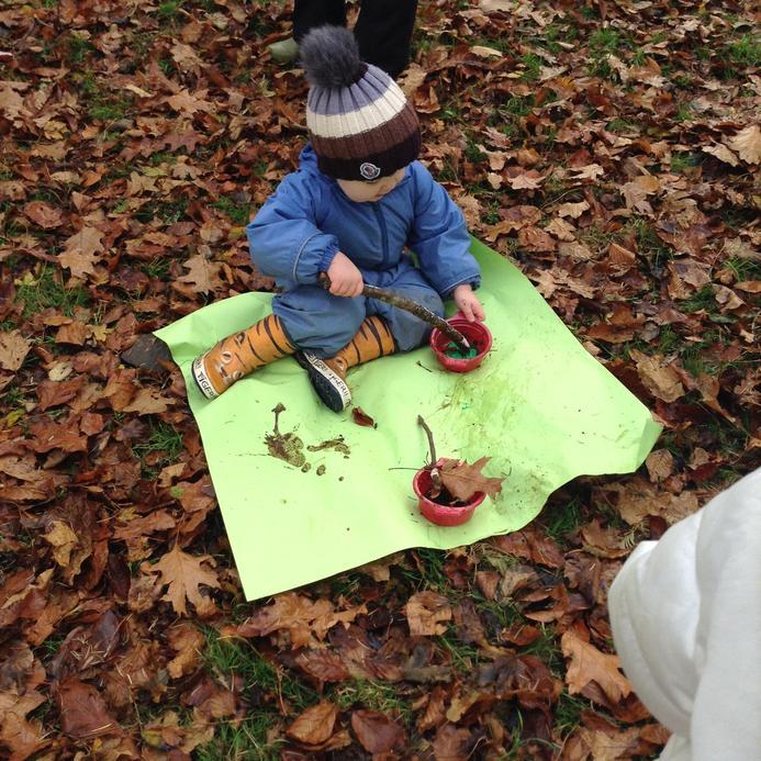 A young child in a blue snowsuit and striped bobble hat sits on a green mat, stirring leaves and mud in red bowls with a stick, surrounded by autumn leaves.