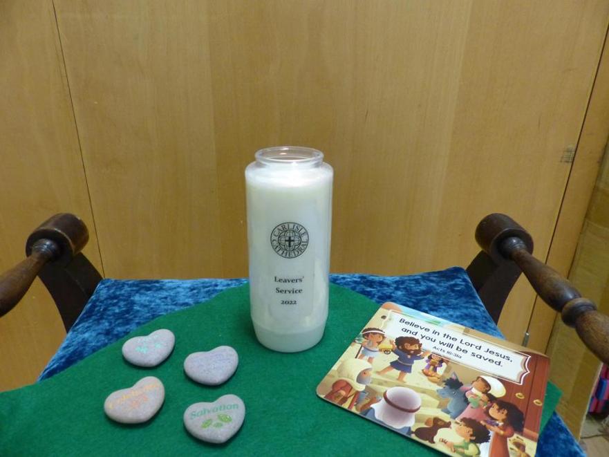 A Carlisle Cathedral Leavers' Service candle, heart-shaped stones, and a scripture card with 'Believe in the Lord Jesus' on a chair.