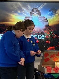 Two schoolgirls in blue uniform observing Remembrance Day, holding poppies in front of a digital memorial background.