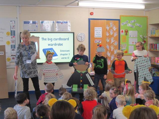 An adult teacher and children present a lesson on virtues using cardboard cutouts in a classroom. Screen shows 'The big cardboard wardrobe' and Colossians 3:12.